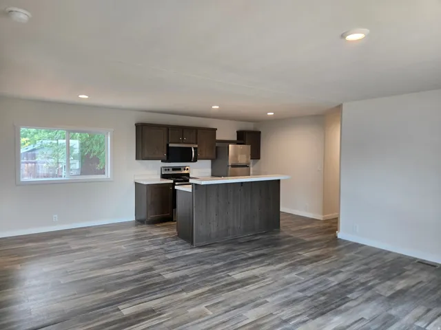 Open-concept living and kitchen area featuring dark cabinetry, stainless steel appliances, a large kitchen island with a breakfast bar and a wood-finish base, recessed ceiling lighting, and wood-finish flooring throughout. Dining nook to the right.