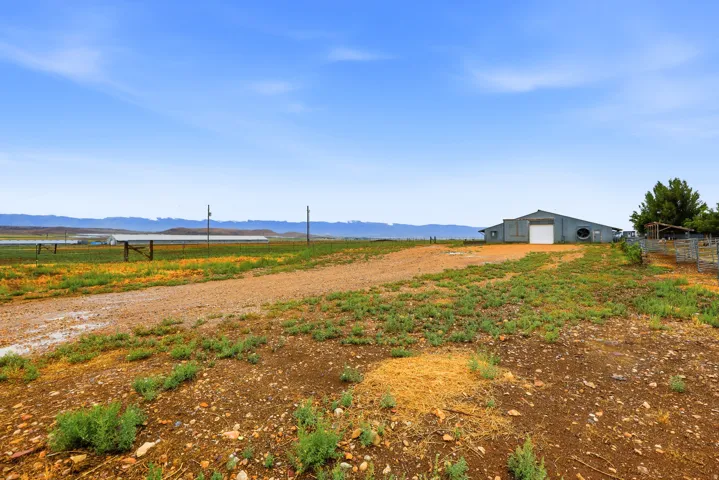 Expansive rural property featuring a large metal barn with a white roll-up door, a gravel pathway, and distant structures under a clear sky