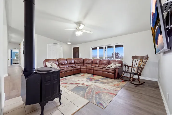 Vaulted ceiling, wood-burning stove with tile surround, wood-finish flooring, ceiling fan, and large windows