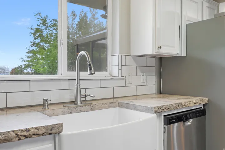 White farmhouse sink with a gooseneck faucet, light-toned countertops, white subway tile backsplash, white upper cabinetry, and a built-in stainless steel dishwasher