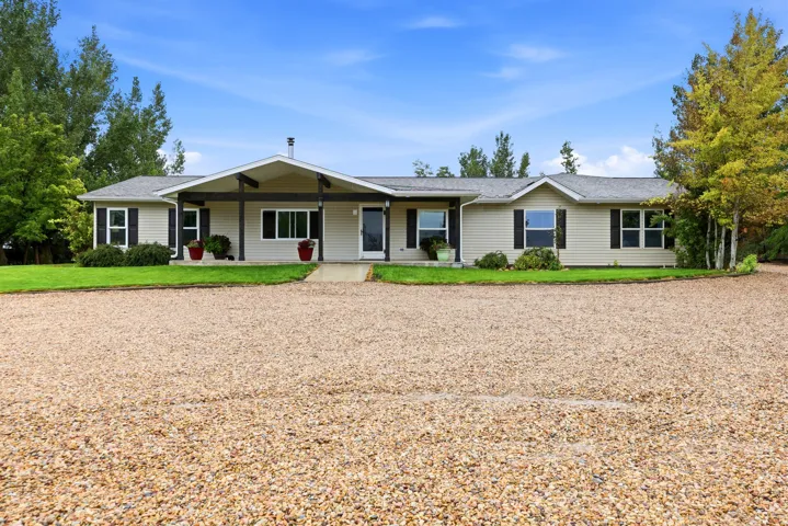 Expansive gravel driveway leading to a residence with a covered entryway, light-colored siding, black window shutters, and a well-maintained lawn