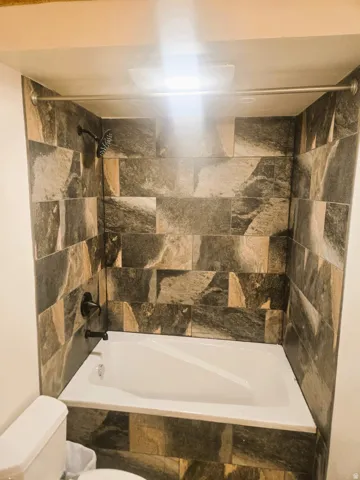 Bathtub with stone-finish tile surround, matte black shower fixtures, and a ceiling-mounted light fixture