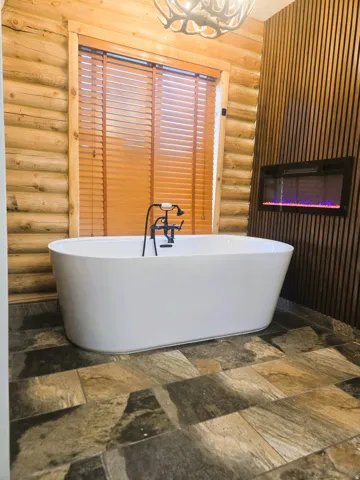 Freestanding soaking tub with a floor-mounted fixture, natural log walls, stone tile flooring, and a built-in electric fireplace within a wood-paneled wall