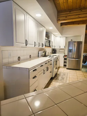 White cabinetry with black hardware, tile countertops, recessed lighting, and exposed wood beams