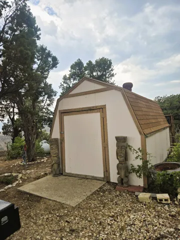 Utility shed featuring a gambrel roof with shingle finish, white siding, and a concrete pad