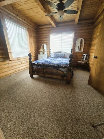 Log-style wall construction, exposed wood beam ceiling, ceiling fan with integrated lighting, window blinds, and neutral-toned carpeting