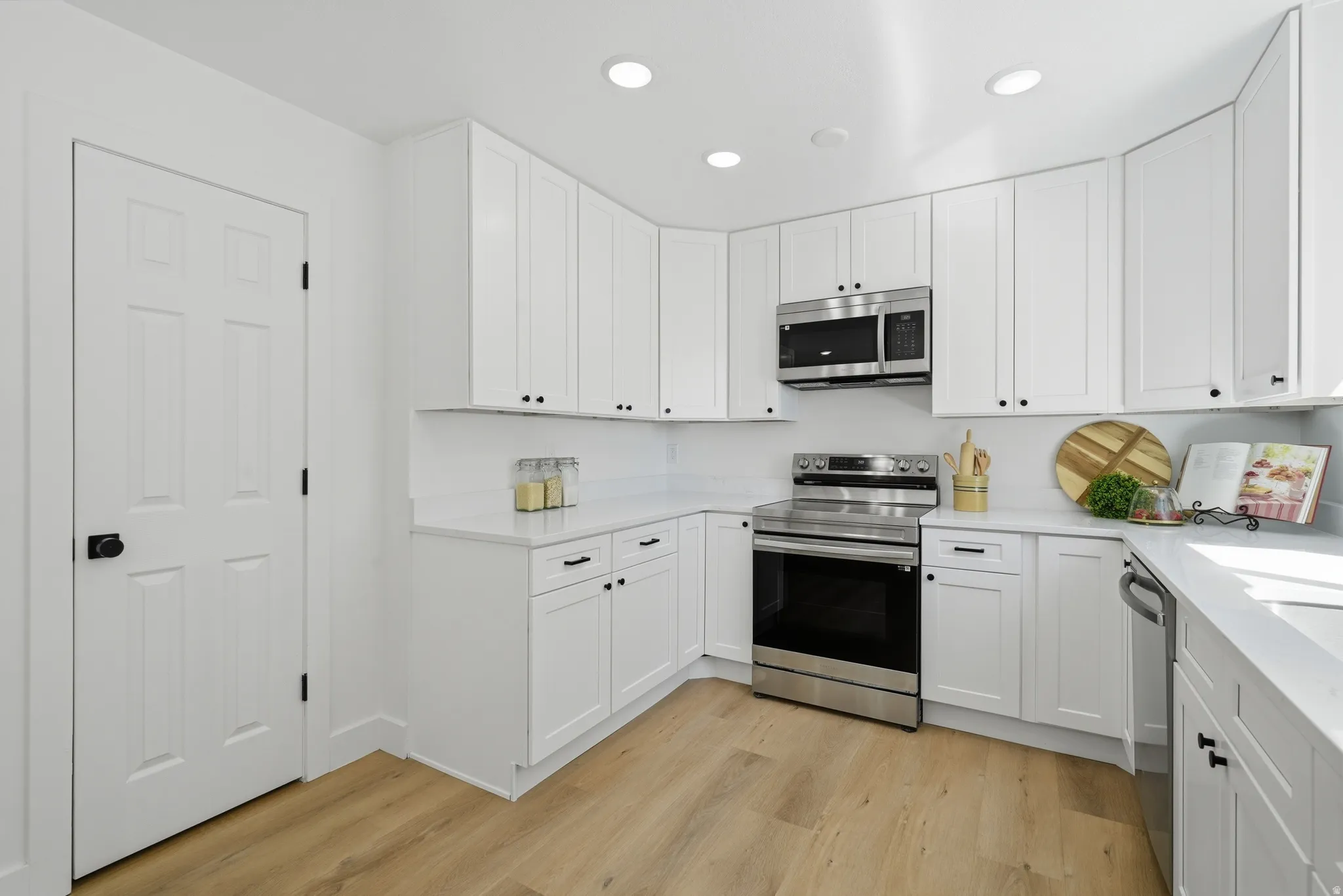 Recessed lighting illuminating white cabinetry, stainless steel appliances, light-toned wood-finish LVP flooring, and quartz countertops with a pantry