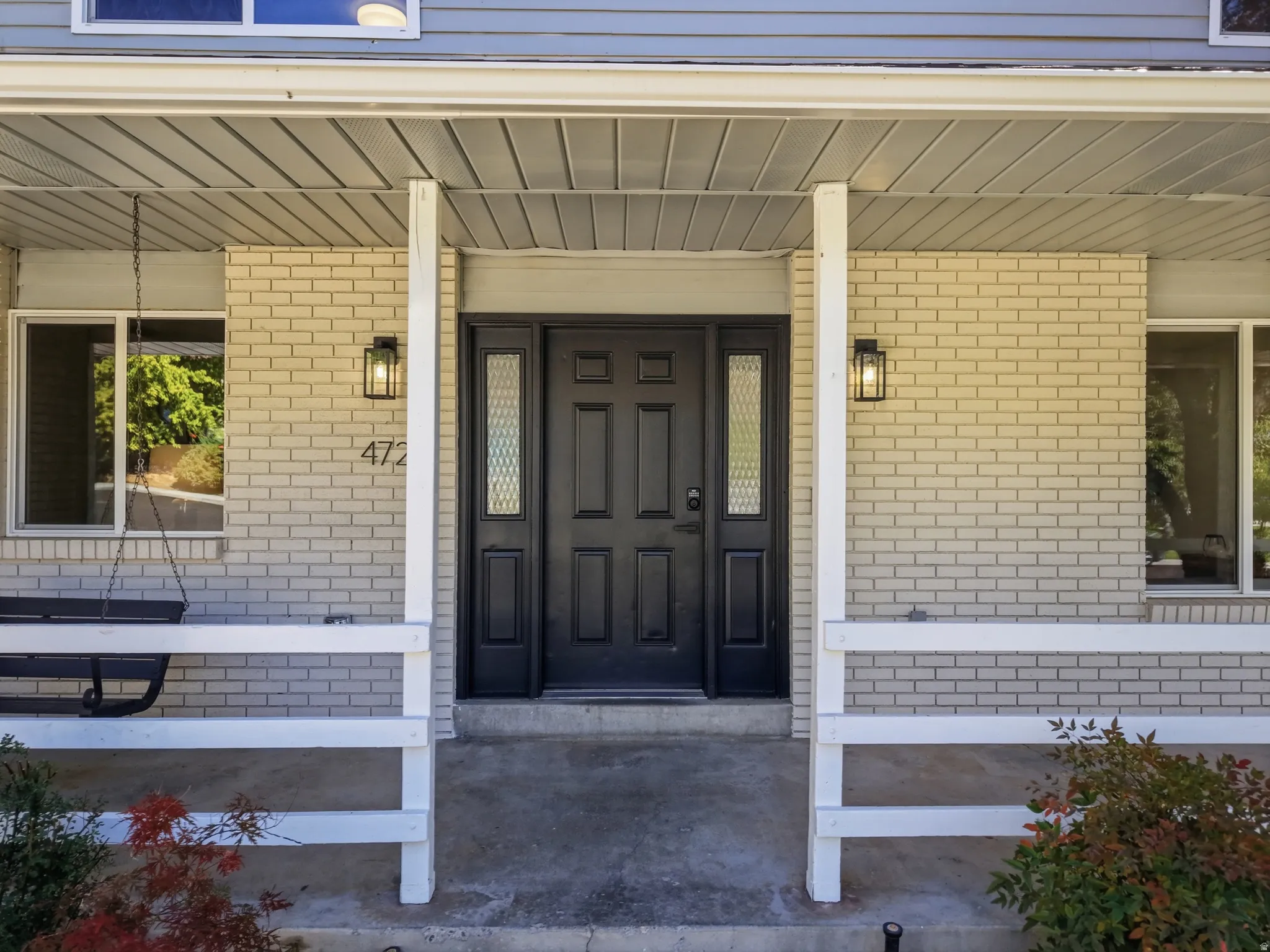 Covered porch and entryway with brick facade, black paneled door with decorative glass sidelights, and two exterior sconces