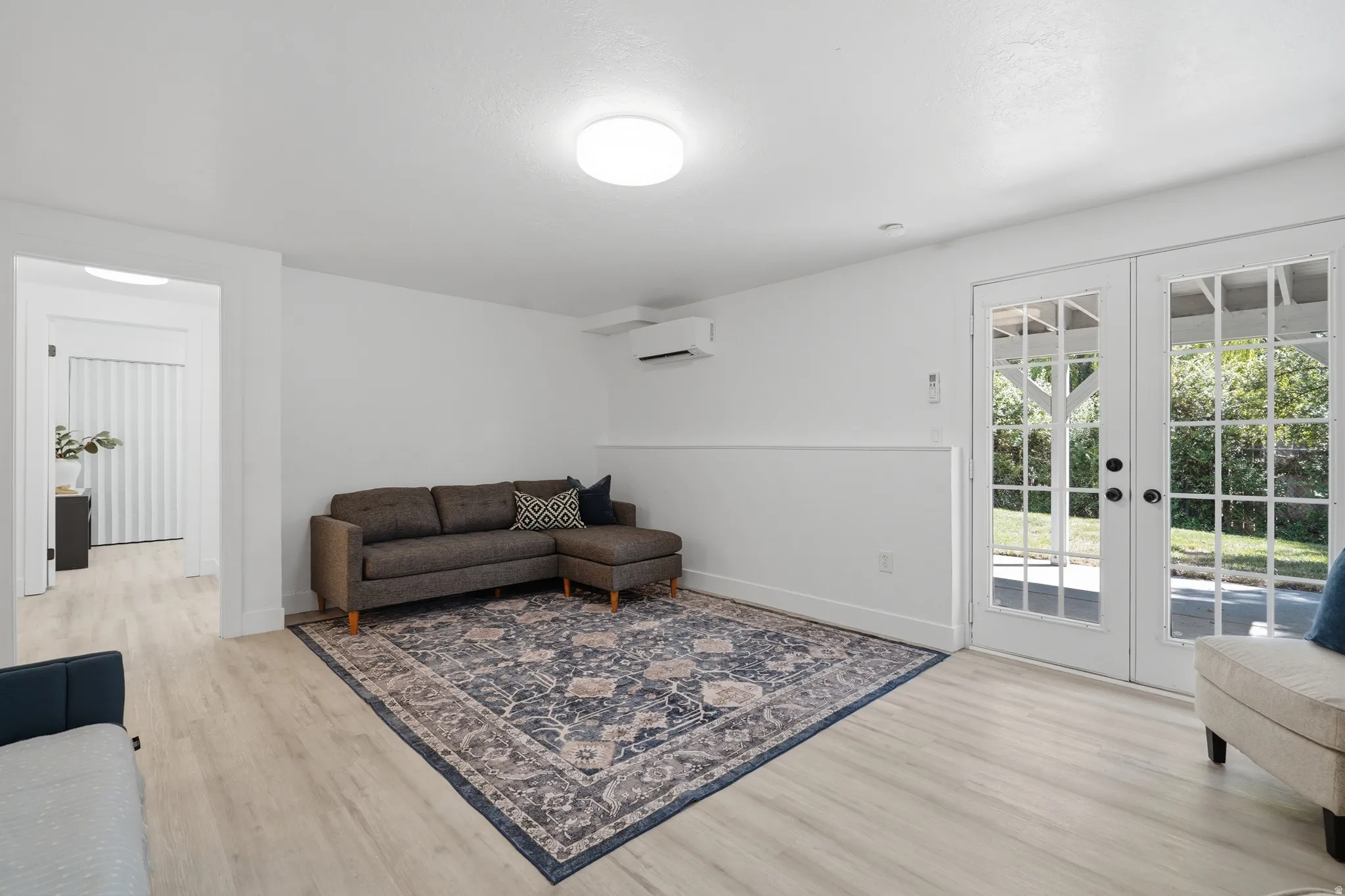 ADU living room with light-toned laminate flooring, white walls, white French doors, and a wall-mounted mini-split unit