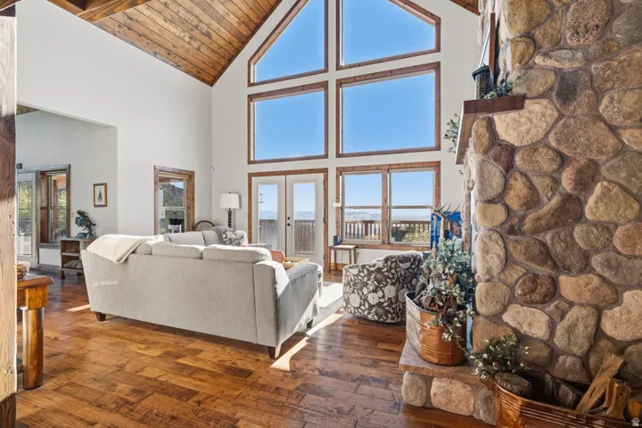Vaulted wood-paneled ceiling, expansive window wall, stone fireplace, wood-finish flooring, and double glass doors leading to a balcony
