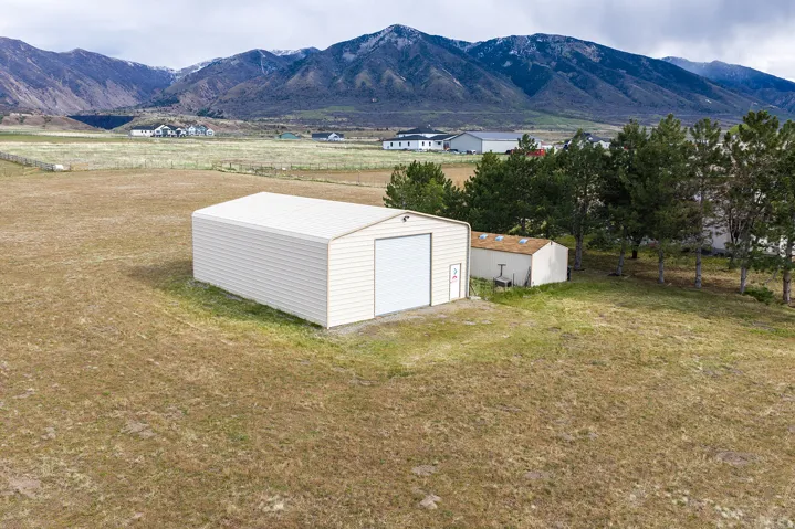Expansive field with a large metal outbuilding featuring a roll-up door, a smaller secondary outbuilding, and mature evergreen trees