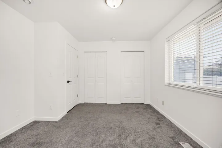 White walls with gray carpet, two bi-fold closet doors, and a window with white blinds