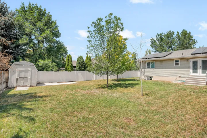 Expansive grassy yard featuring a white privacy fence, mature trees, a storage shed, and a concrete patio