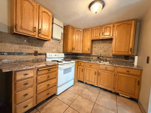 Kitchen featuring wood-finish cabinetry, tile flooring, brick-patterned backsplash with decorative accents, and a built-in stainless steel sink