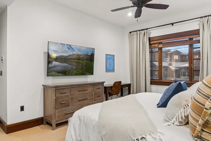 Wall-mounted television with built-in desk, wood-framed window, and ceiling fan with integrated lighting