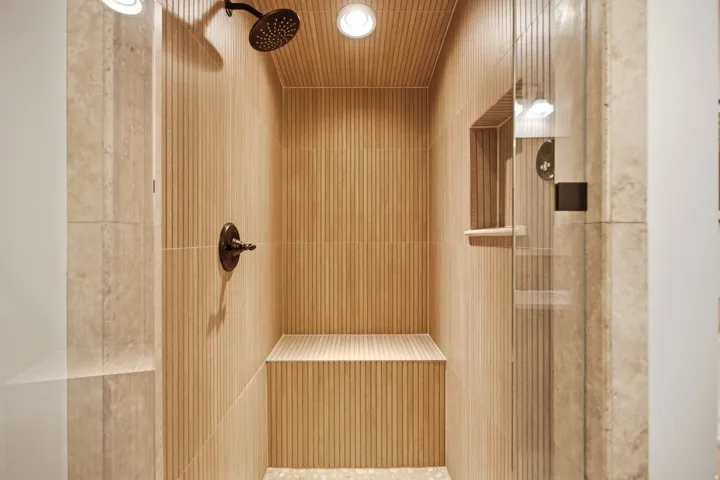 Walk-in shower featuring vertically ribbed tile, a built-in bench, a recessed niche, a dark bronze rainfall showerhead, and fixed glass panels