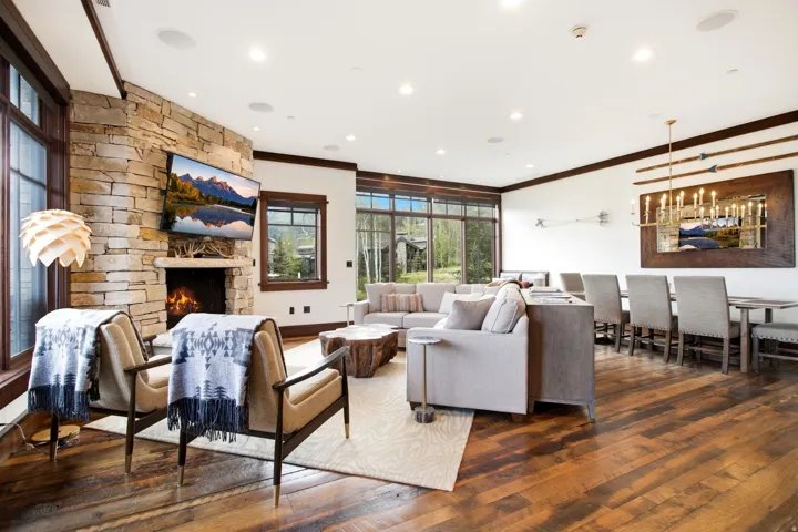 Spacious living area featuring a stone-clad fireplace, wood-finish flooring, large windows, and a dining area with a chandelier