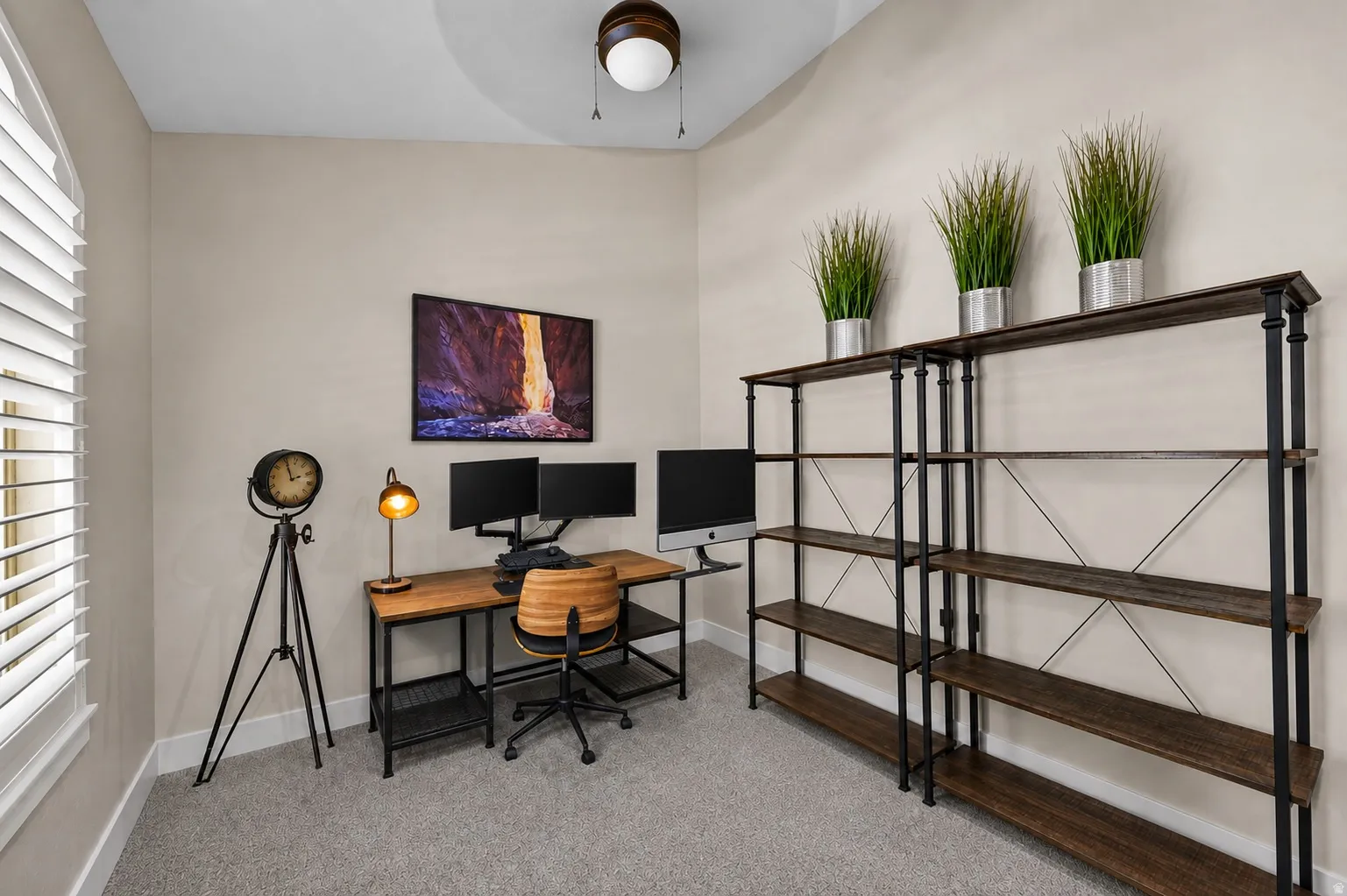 Arched window with plantation shutters, ceiling fan with integrated lighting, built-in shelving, wood-finish desk, and carpeted flooring