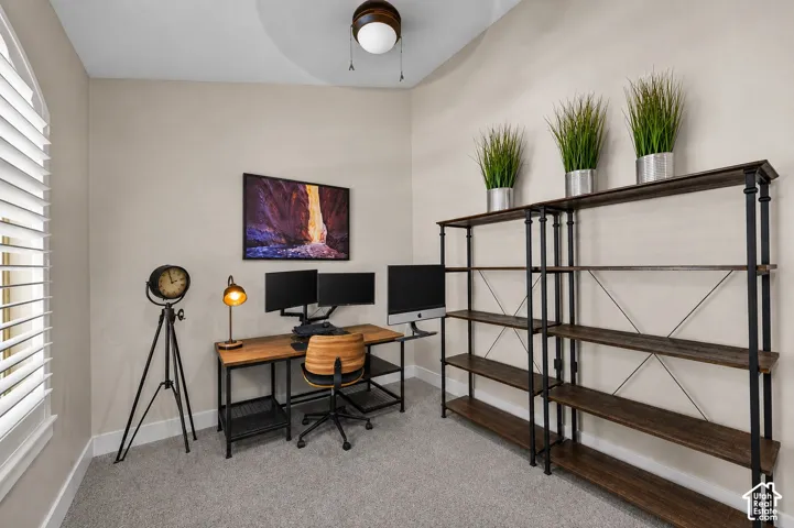 Arched window with plantation shutters, ceiling fan with integrated lighting, built-in shelving, wood-finish desk, and carpeted flooring