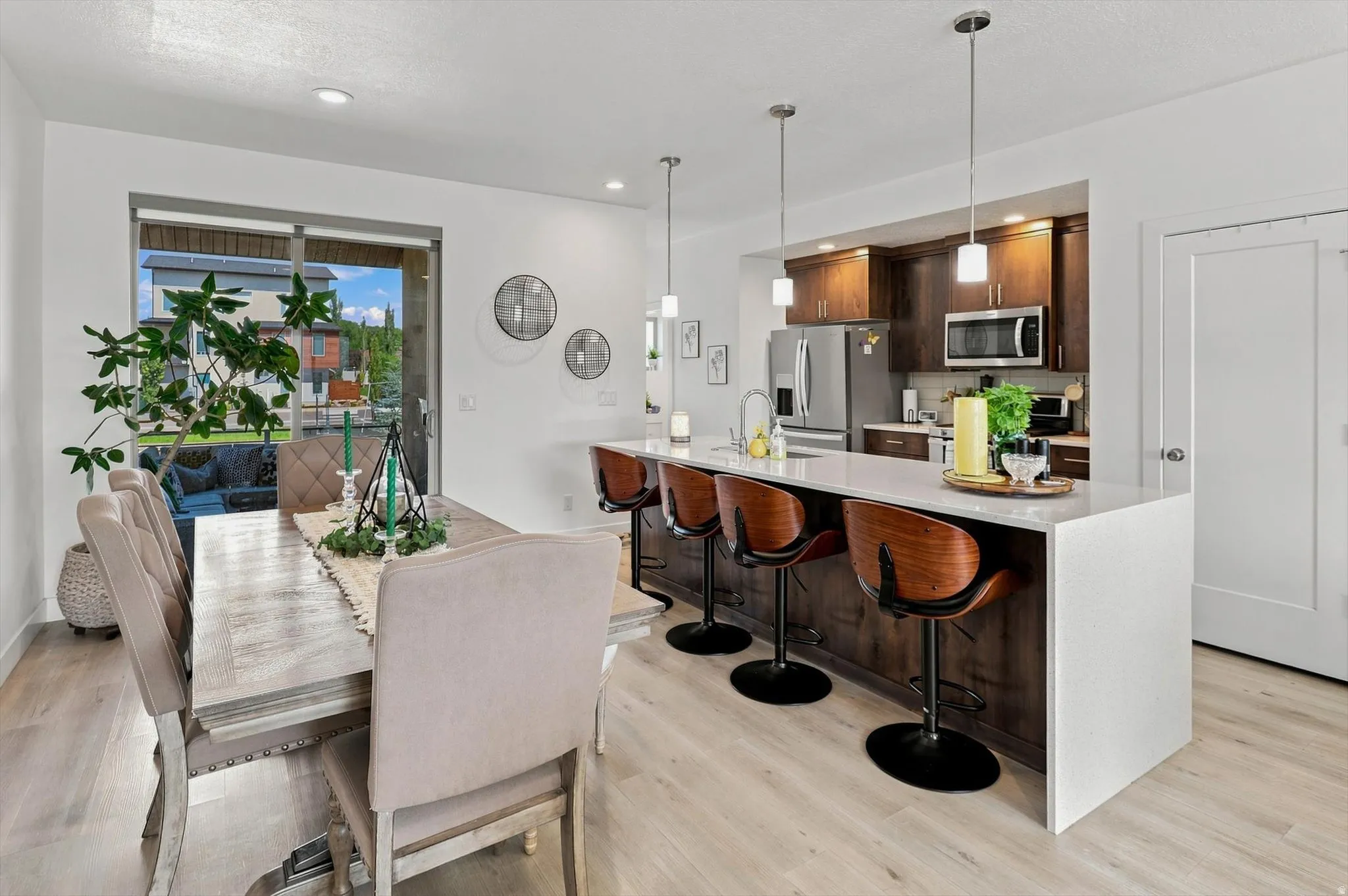 Open-concept kitchen and dining area featuring a waterfall island with a light-toned countertop, dark wood-finish cabinetry, stainless steel appliances, and pendant lighting
