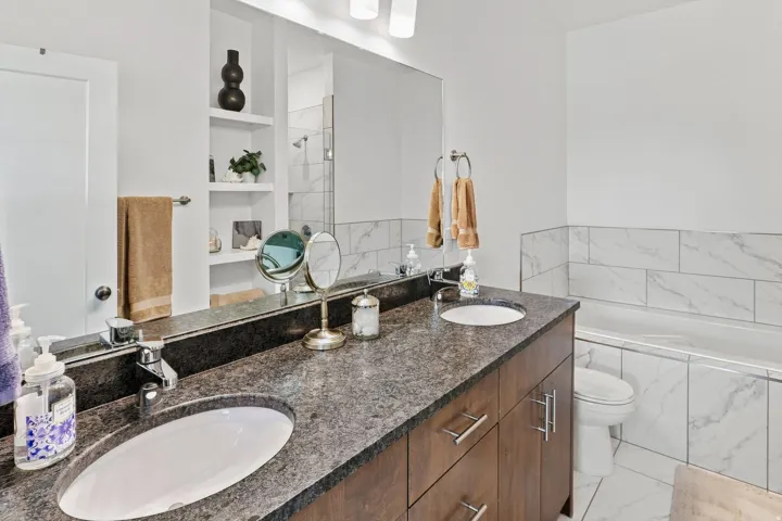 Dual vanity with undermount sinks and dark stone countertop, built-in shelving, white subway tile surround, and an integrated bathtub