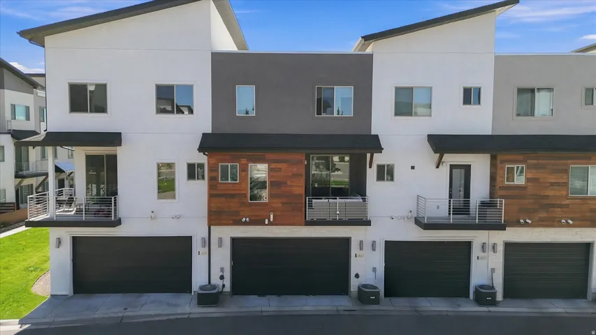 Contemporary townhome exterior featuring an attached two-car garage, private balcony with metal railing, and a mix of white stucco, dark gray siding, and wood-panel accents