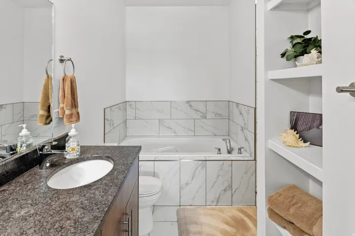 Built-in bathtub with subway tile surround, vanity with dark stone countertop, undermount sink, and integrated shelving