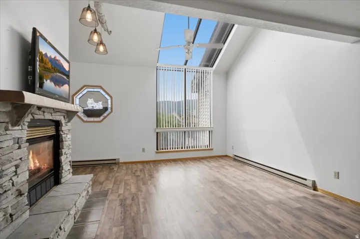 Vaulted ceiling, stone-clad fireplace with hearth, wood-finish flooring, full-height window with vertical blinds, and a skylight