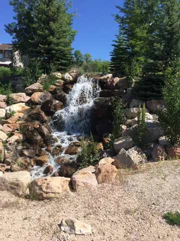 Rock waterfall feature with natural stone landscaping, surrounded by mature and evergreen trees, It is the gateway to extensive walking trails