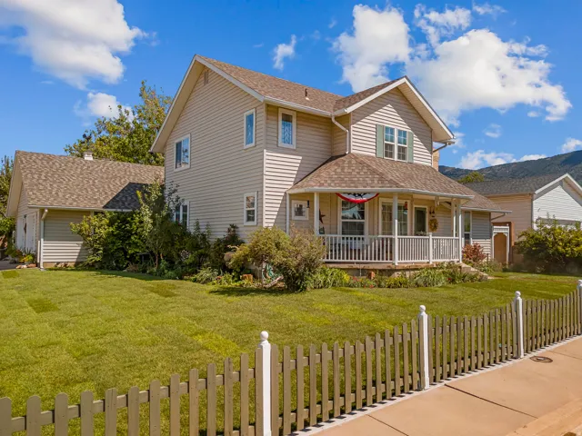 Two-story residence with light-colored siding, a covered front porch featuring white railings, a detached structure with a pitched roof, a front lawn, and a picket fence