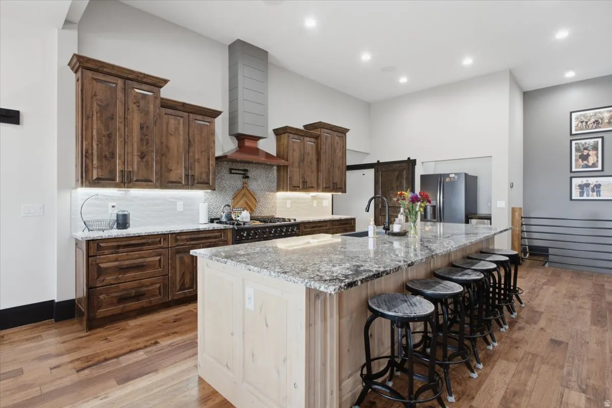 Spacious kitchen featuring wood-finish flooring, an expansive island with a stone countertop, dark wood cabinetry, a range with a copper-tone hood, and recessed lighting