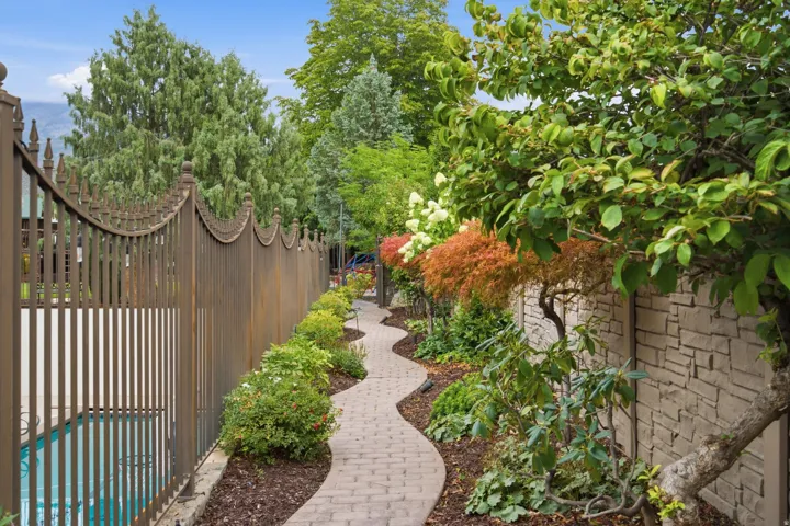 Winding brick pathway bordered by diverse plantings, decorative metal fencing, and a stone-clad wall