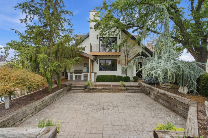 Exterior with white siding and black trim, featuring a covered patio with an outdoor kitchen, second-story balcony with metal railing, and a stamped concrete patio with wood-tie retaining walls