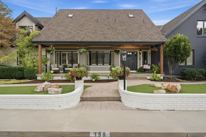 Exterior featuring a covered porch with exposed beams, front yard with artificial turf, white brick retaining wall, and a stamped concrete pathway