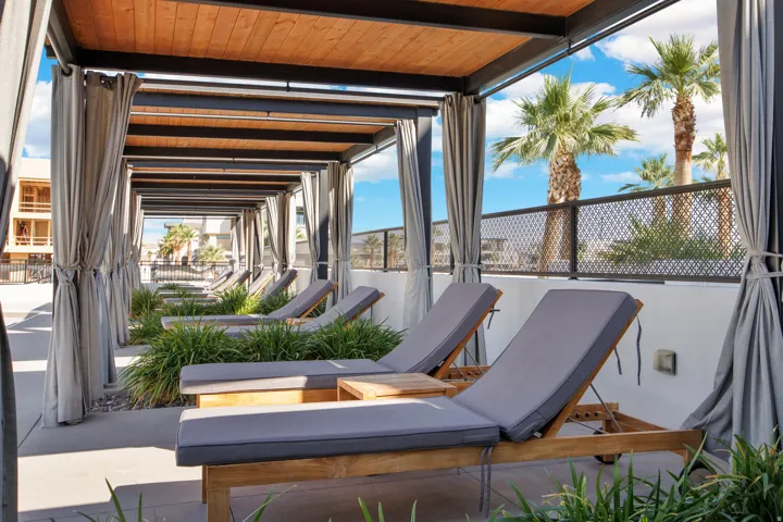 Covered lounge area featuring wood-finish ceiling beams, gray upholstered chaise lounges with wood frames, sheer gray curtain panels, mature palm trees, and a metal mesh safety railing overlooking the surrounding landscape