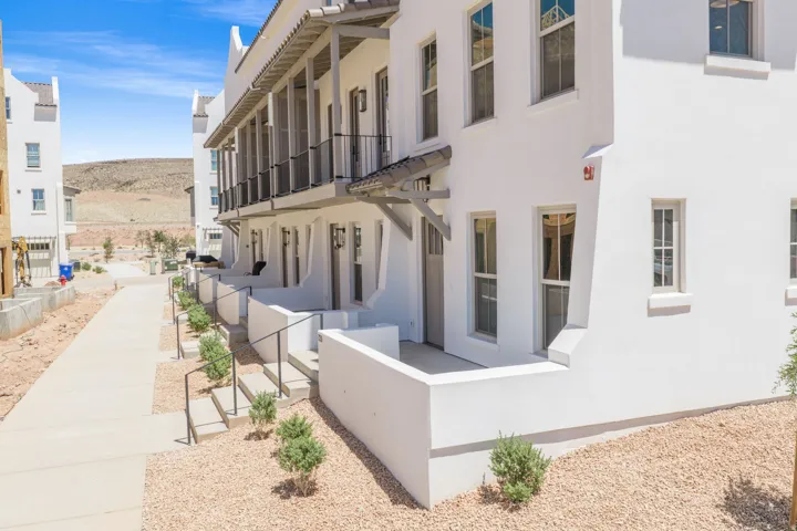 White stucco exterior with multiple windows, individual patio entrances, upper-level balconies with black railings, and a concrete pathway with landscaped gravel areas