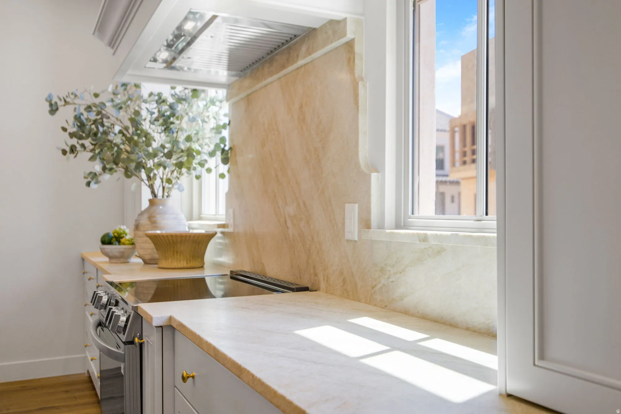 Built-in stainless steel range, light-toned stone countertops, matching stone backsplash, under-cabinet lighting, and white cabinetry with brass-finish hardware