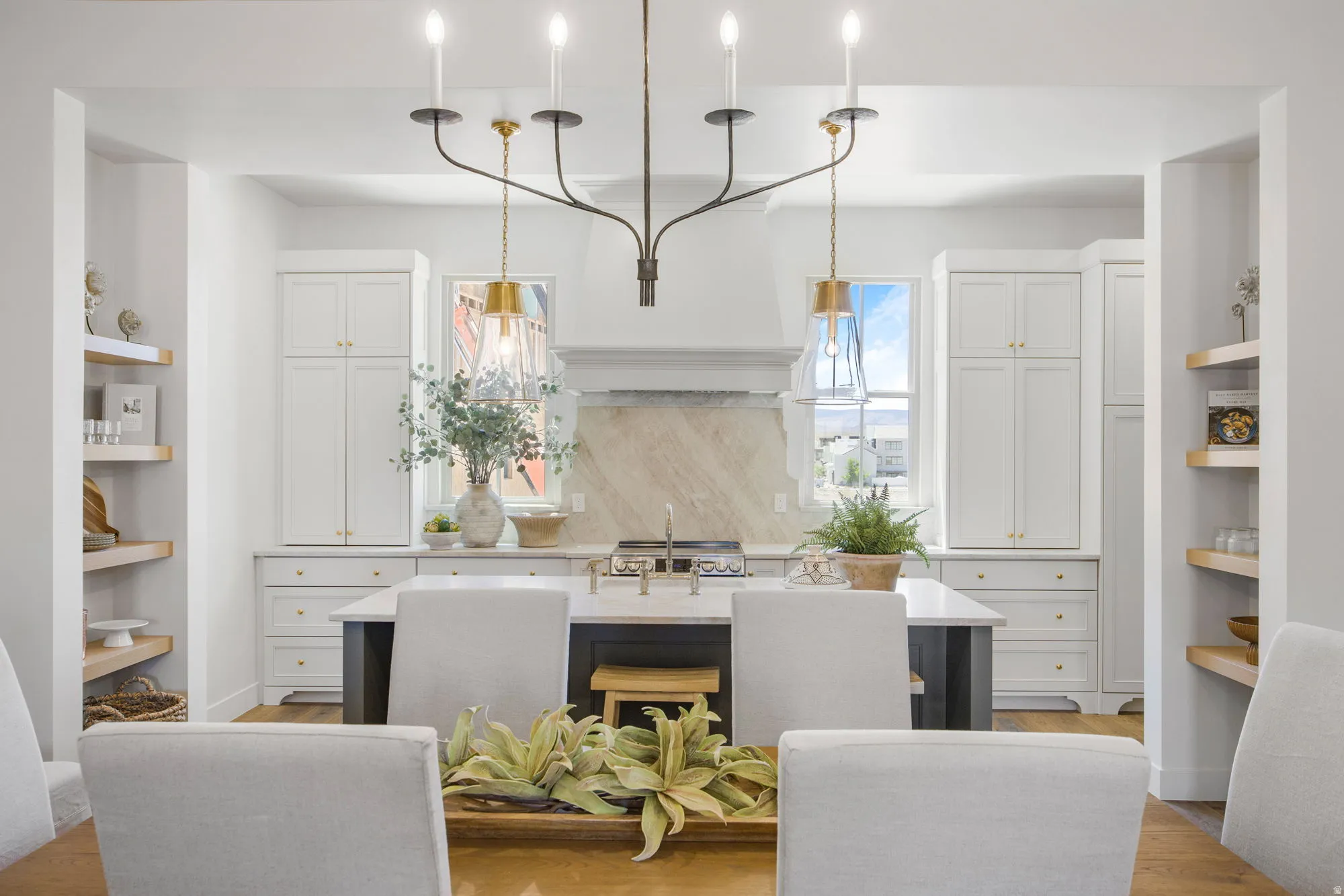 White shaker-style cabinetry with gold-tone hardware, a center island with cooktop, a decorative range hood, and light wood-finish open shelving