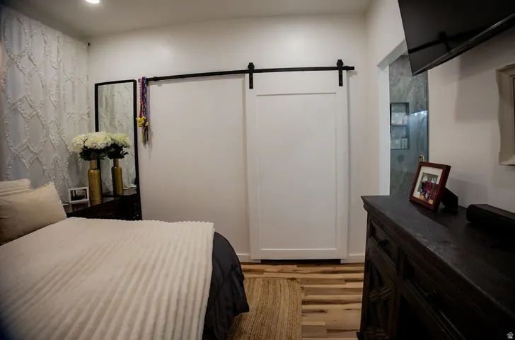 White paneled barn door on a black track, light wood-finish flooring, recessed ceiling lighting, and a partial view of a walk-in shower with glass enclosure