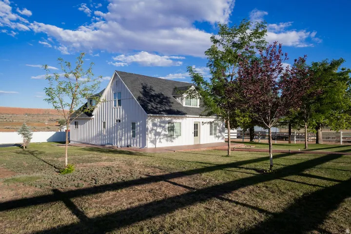 White board and batten siding, dark shingle roof, dormer windows, mature trees, and a white perimeter fence