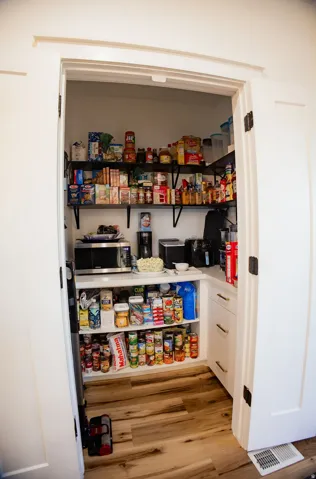 Walk-in pantry featuring wood-finish flooring, white cabinetry, a white countertop, and dark-finish shelving
