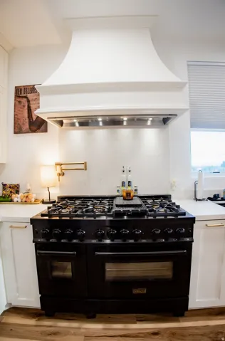 White range hood with integrated lighting, large black professional-style range, white subway tile backsplash, white shaker-style cabinetry, and wood-finish flooring