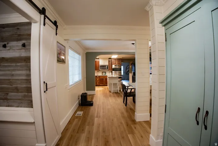 Wood-finish flooring, white barn door on black hardware, shiplap wall treatment, crown molding, and window with blinds