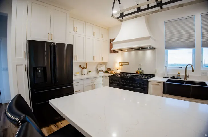 White shaker cabinetry, black stainless steel appliances, large kitchen island with light-toned countertop, range hood, and double basin sink