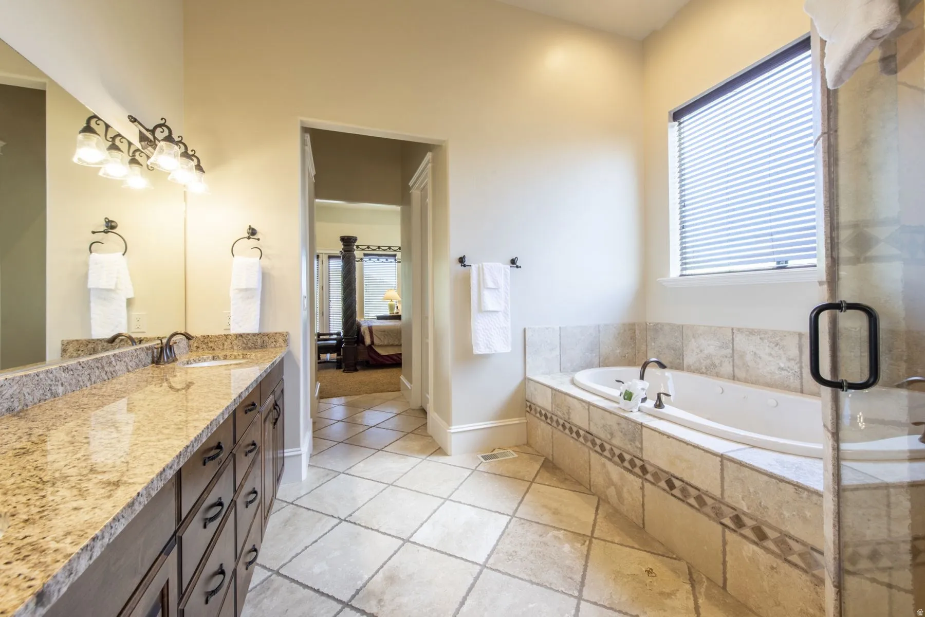 Dual vanity with granite countertop, built-in soaking tub, tiled flooring, glass-enclosed shower, and a window with privacy blinds