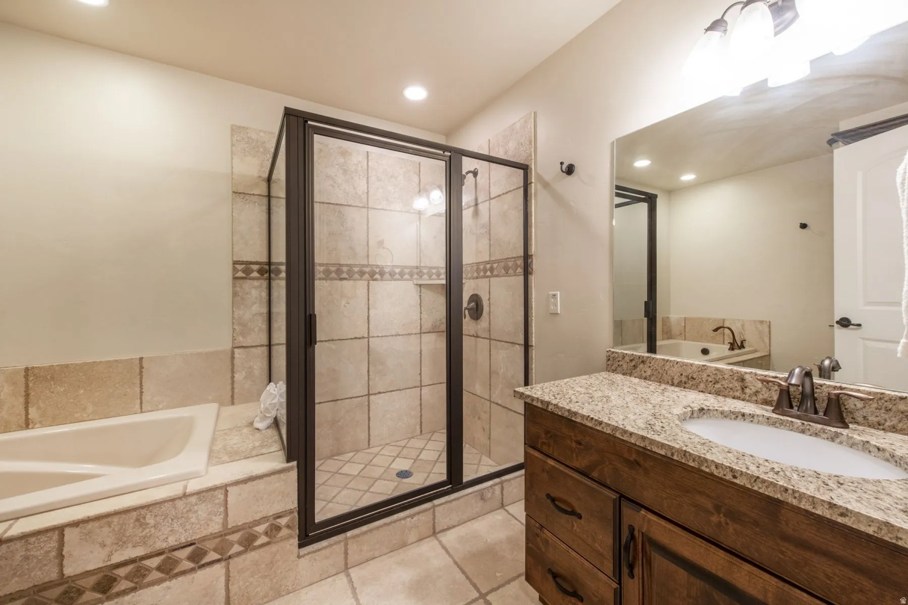 Spa-inspired primary bathroom featuring a frameless glass-enclosed shower with tile surround, dual-sink vanity with granite countertop and wood cabinetry, recessed lighting, and a soaking tub positioned along the tiled wall