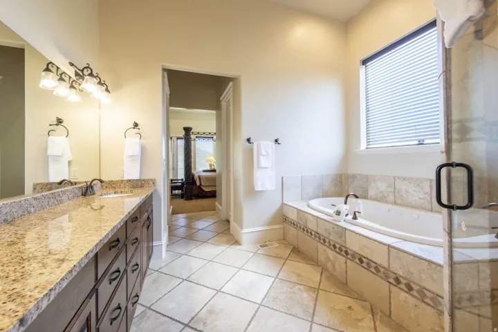 Dual vanity with granite countertop, built-in soaking tub, tiled flooring, glass-enclosed shower, and a window with privacy blinds