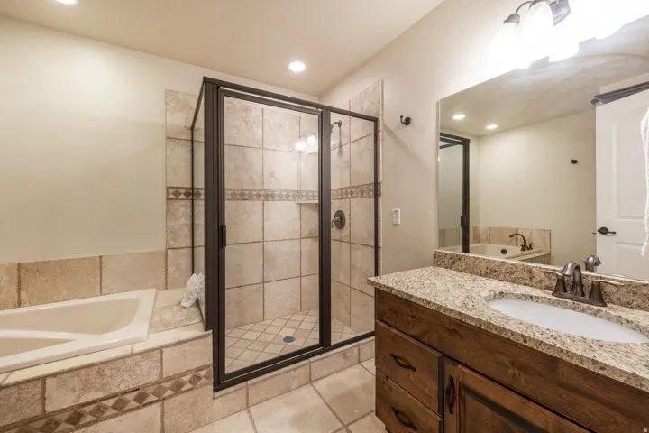 Spa-inspired primary bathroom featuring a frameless glass-enclosed shower with tile surround, dual-sink vanity with granite countertop and wood cabinetry, recessed lighting, and a soaking tub positioned along the tiled wall