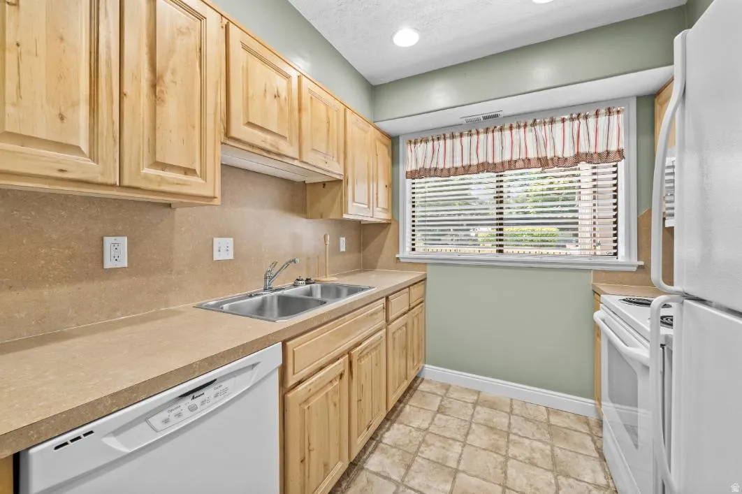 Kitchen featuring light wood cabinetry, a double basin stainless steel sink, light-toned countertops, and a white dishwasher