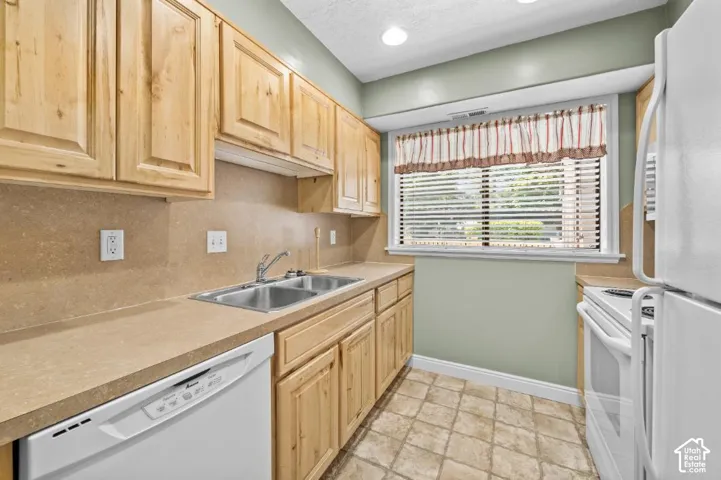 Kitchen featuring light wood cabinetry, a double basin stainless steel sink, light-toned countertops, and a white dishwasher